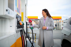 Woman in a gray coat pumping gas at a station, smiling as she refuels a silver car.