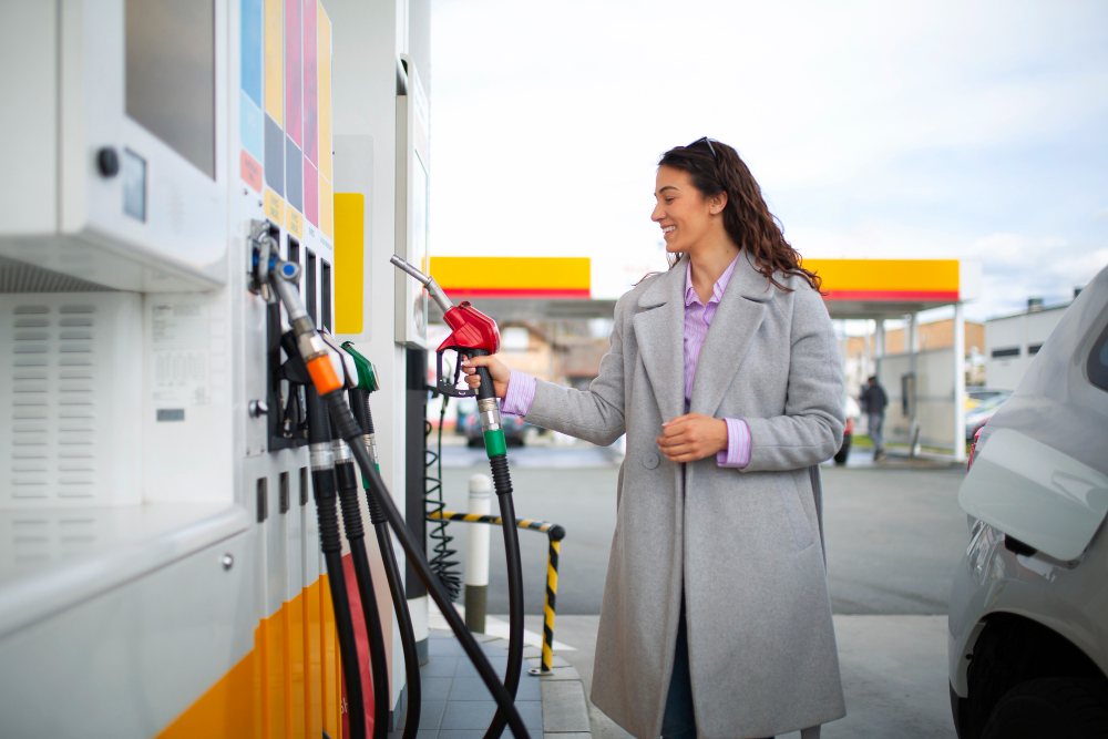 Woman in a gray coat pumping gas at a station, smiling as she refuels a silver car.